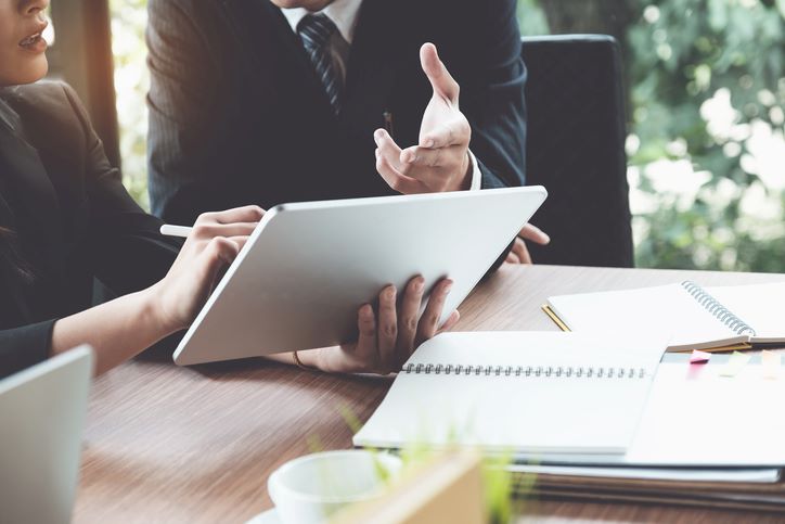 A professional discussion with two people at a desk, one of whom is using a tablet.